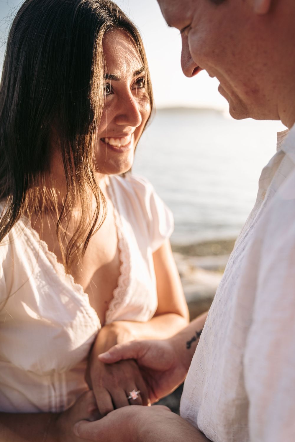 Smiling woman in white dress holds hands with partner near sparkling ocean water in romantic seaside moment.