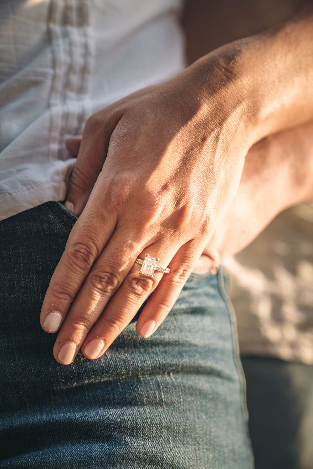 Close-up of an engagement ring on a woman's hand resting on a man's lap in golden sunlight.