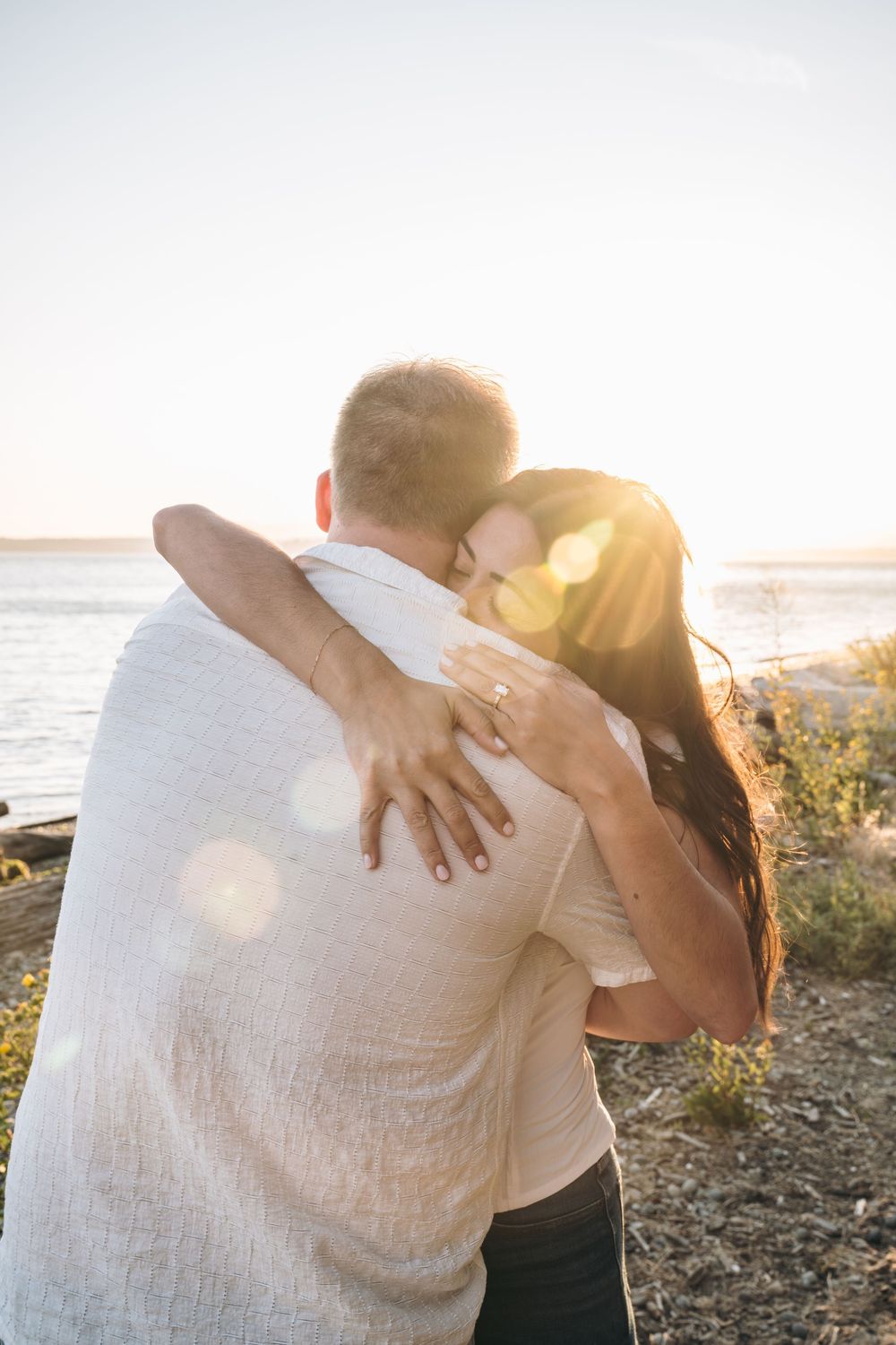 Couple embracing at sunset near the ocean, woman showing off her engagement ring on his back.