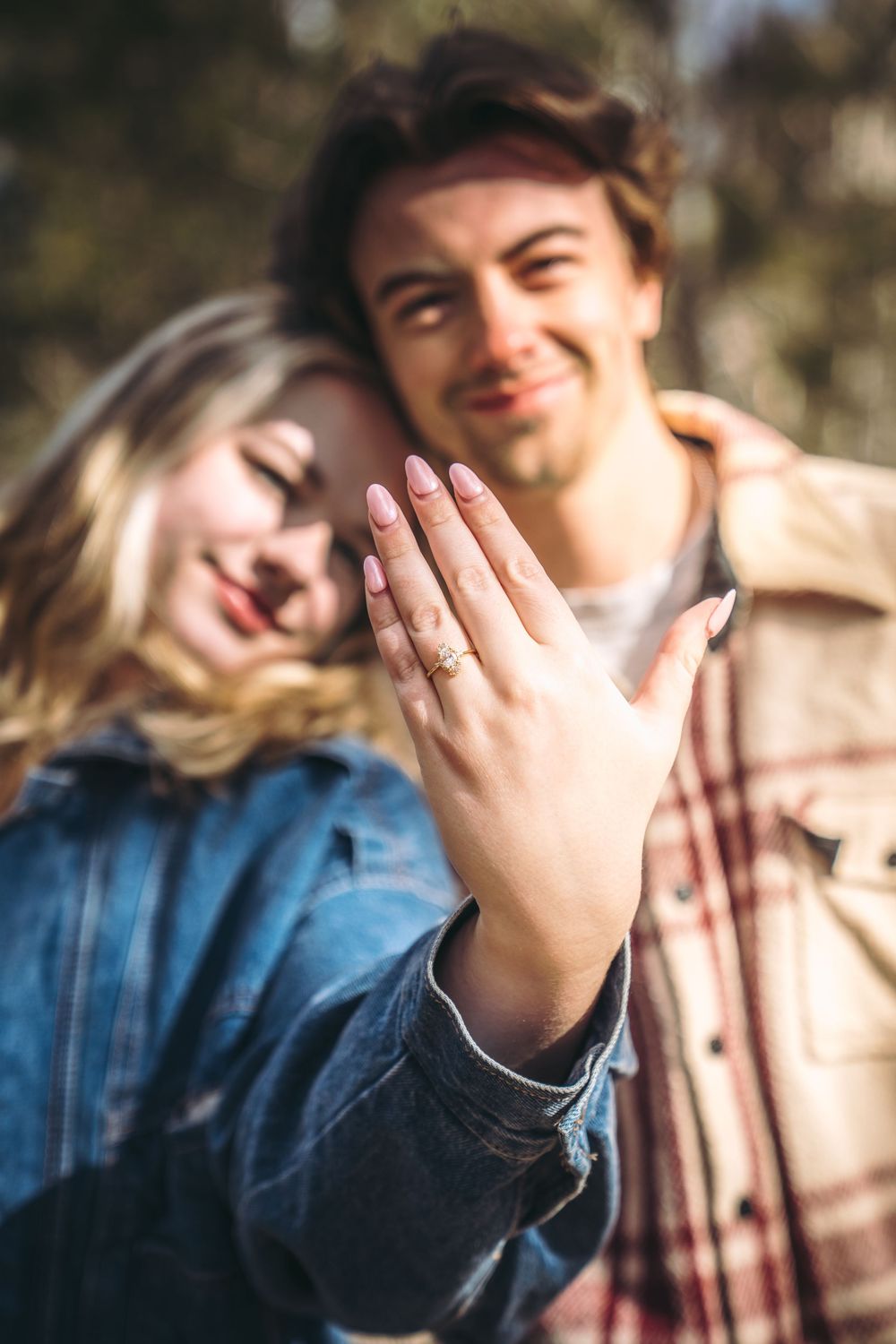 Happy couple showing off engagement ring outdoors, woman displaying diamond ring on hand while leaning on smiling man.