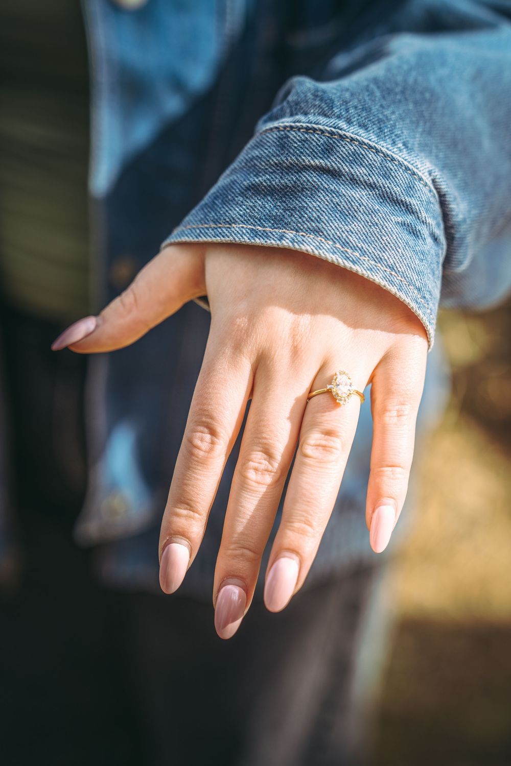 Woman displaying a gold engagement ring with gemstone on ring finger, wearing a denim jacket, with neutral pink manicure.