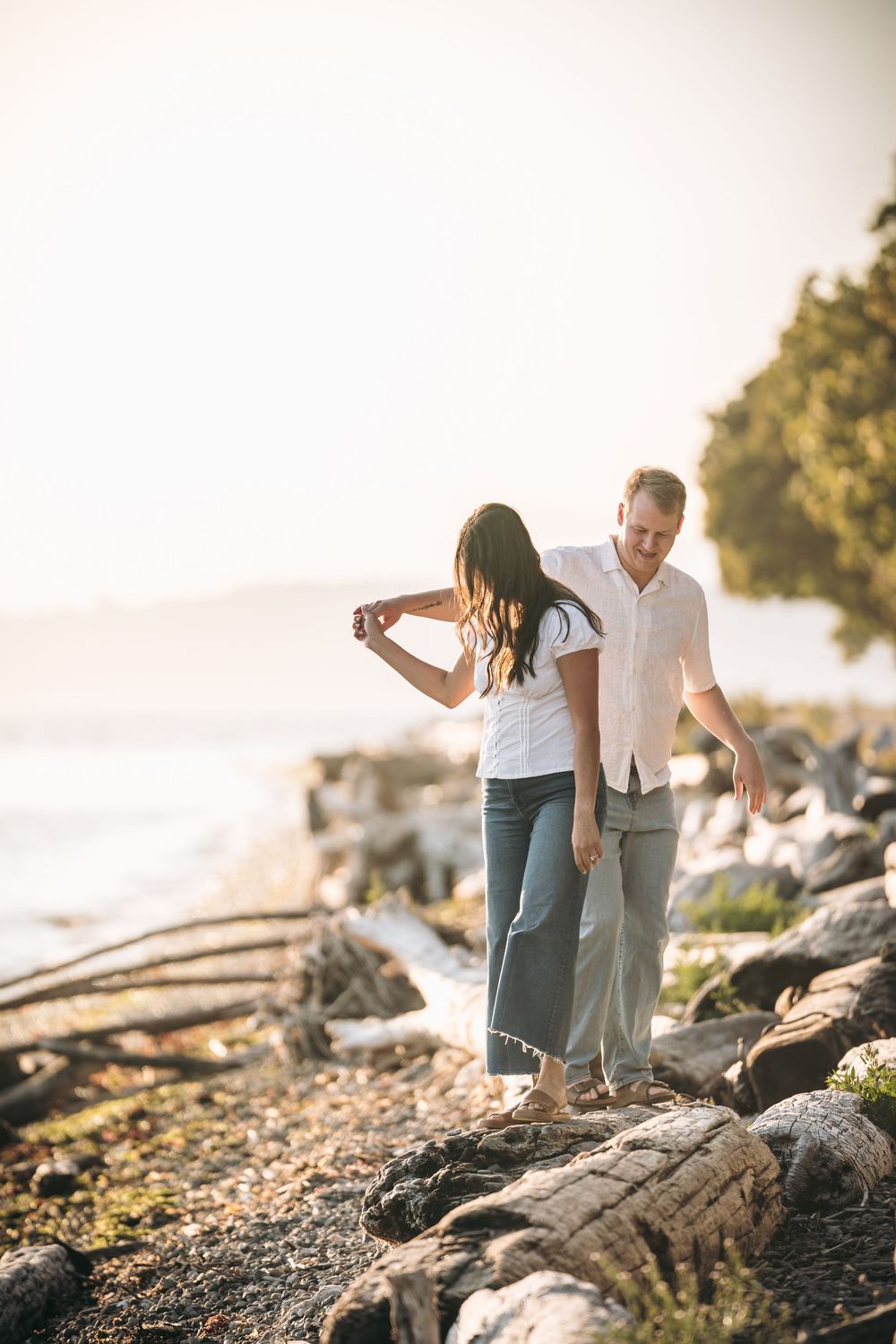 Couple walking hand in hand on rocky shoreline at golden hour, woman in wide-leg pants, man in linen shirt.