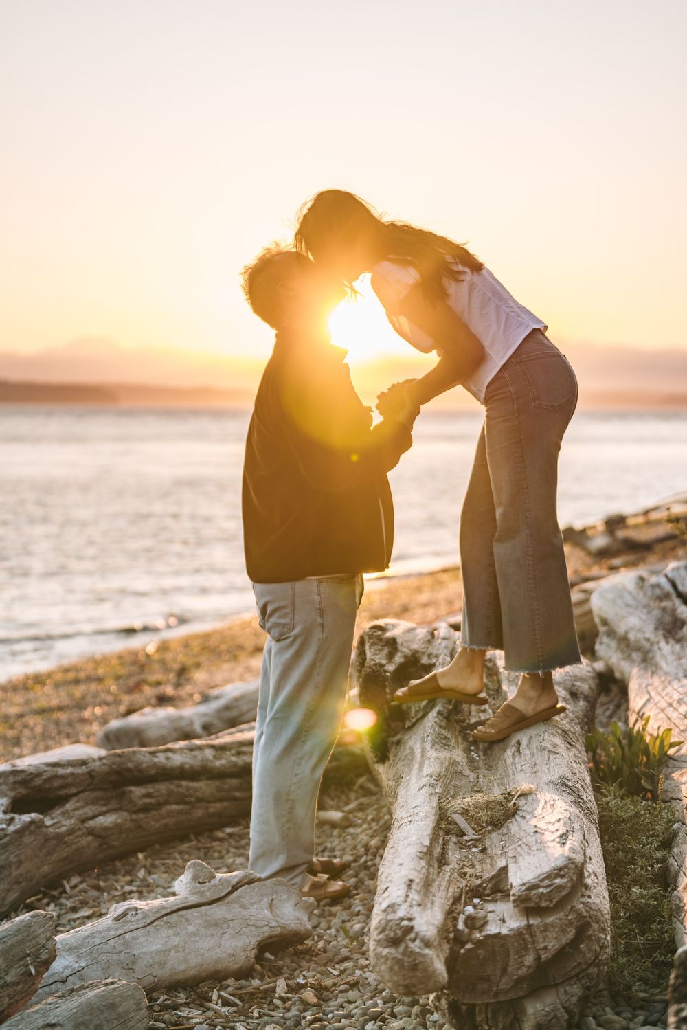 Couple sharing a romantic kiss on driftwood at golden hour sunset by the ocean beach.