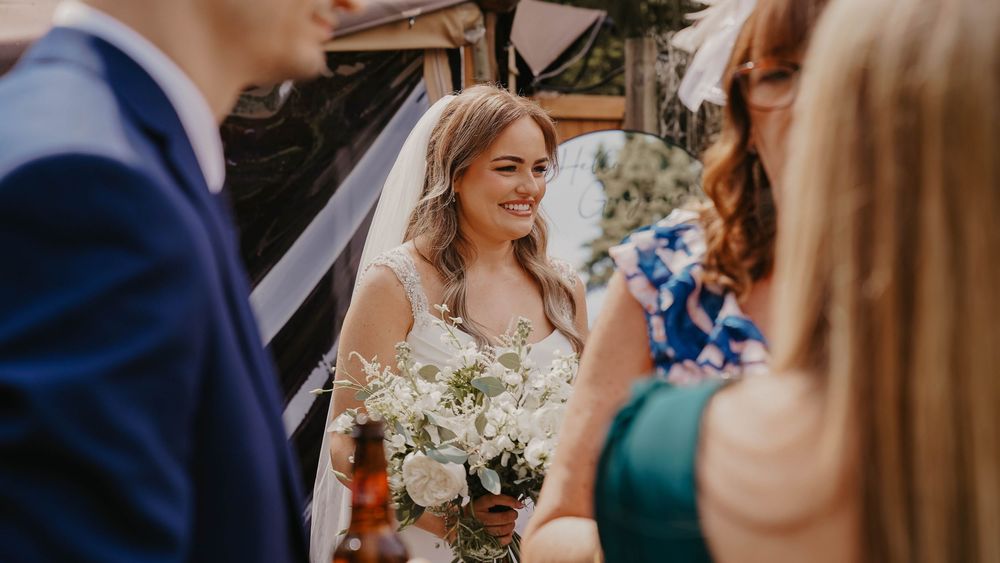 Smiling bride holding white floral bouquet greeting guests outdoors at a wedding ceremony.