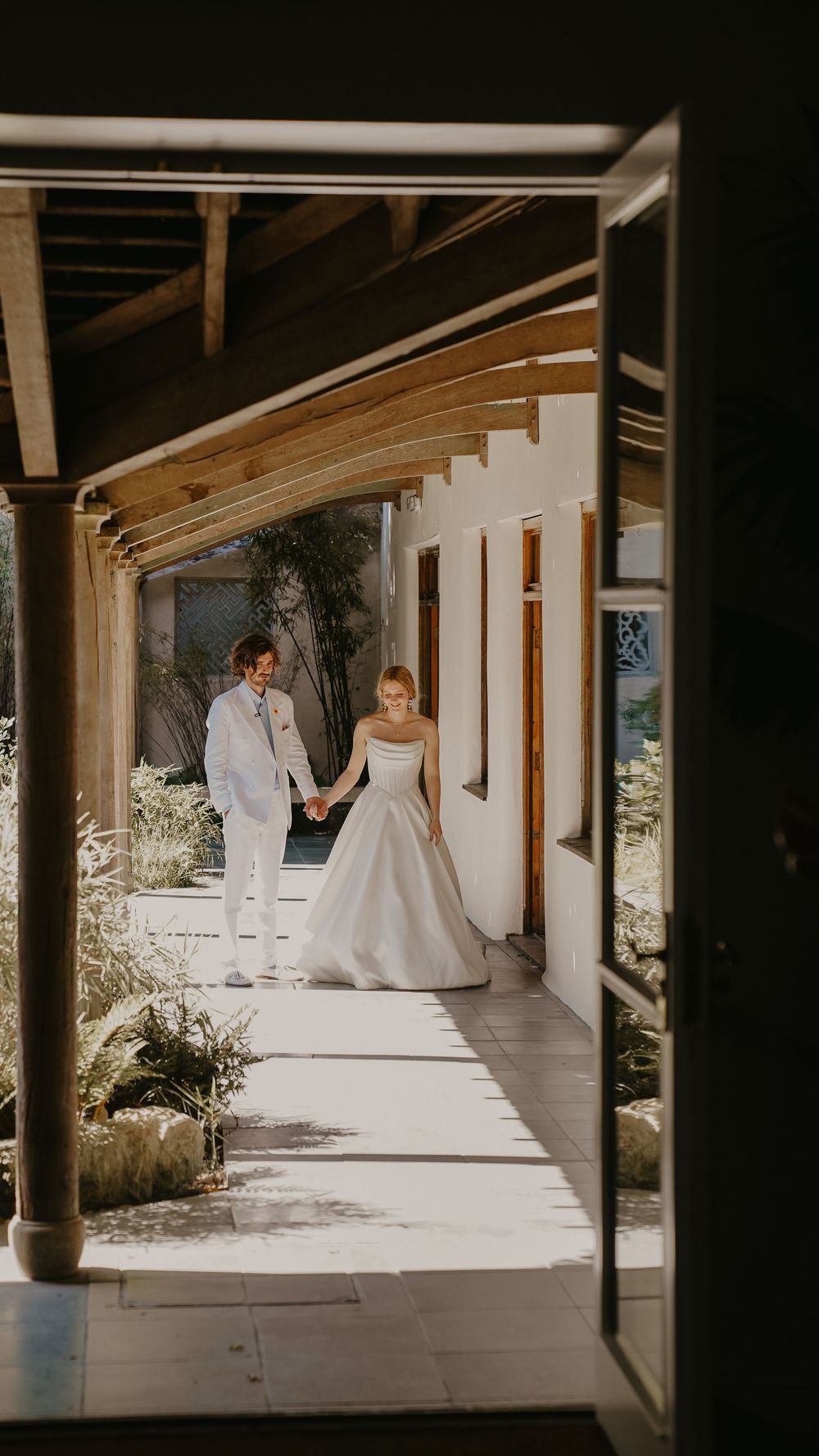 Bride and groom holding hands walking along sunlit outdoor corridor of elegant white venue on wedding day.