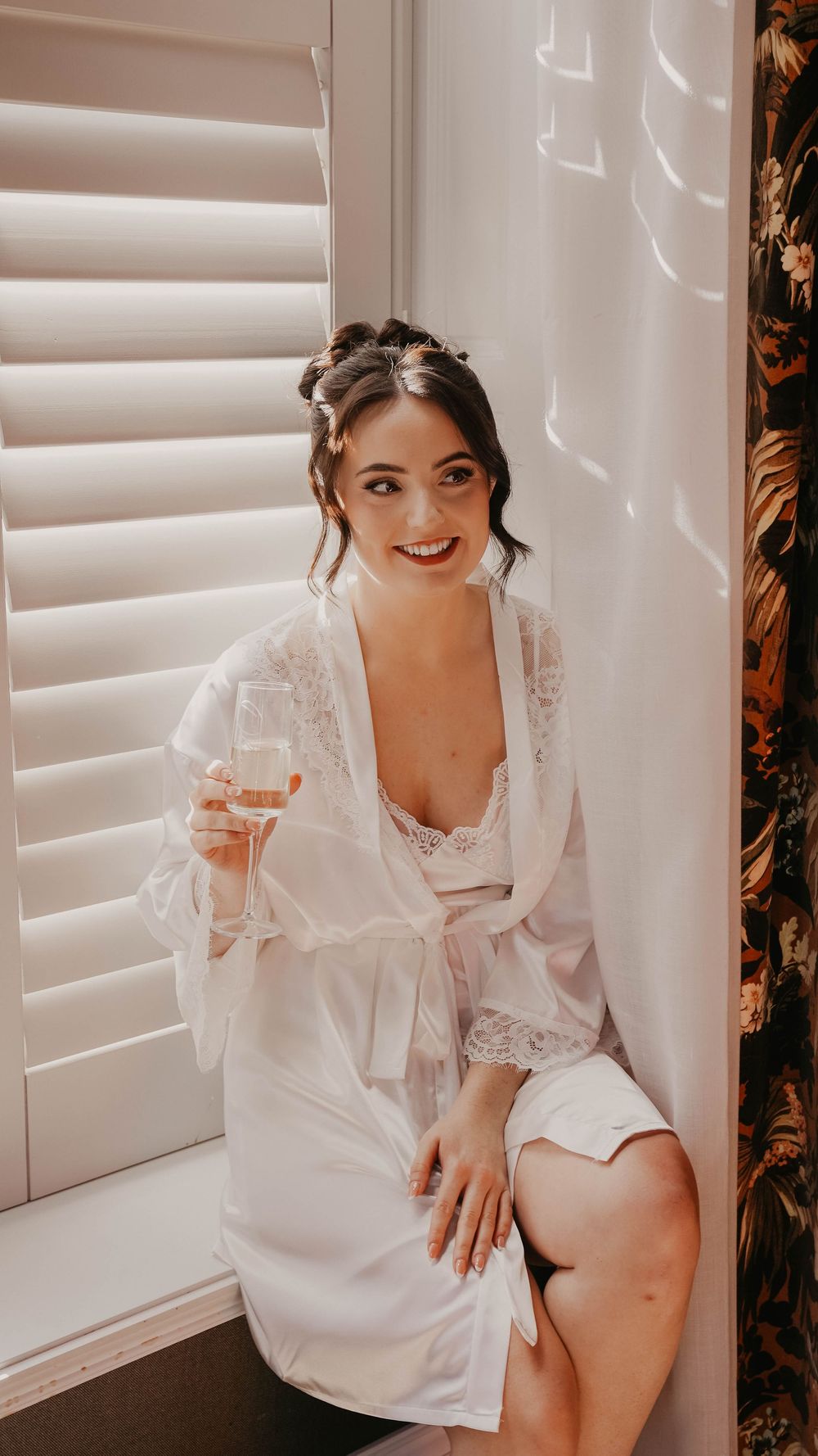 Smiling bride in white lace robe holding champagne glass, sitting by window with shutters on wedding morning.