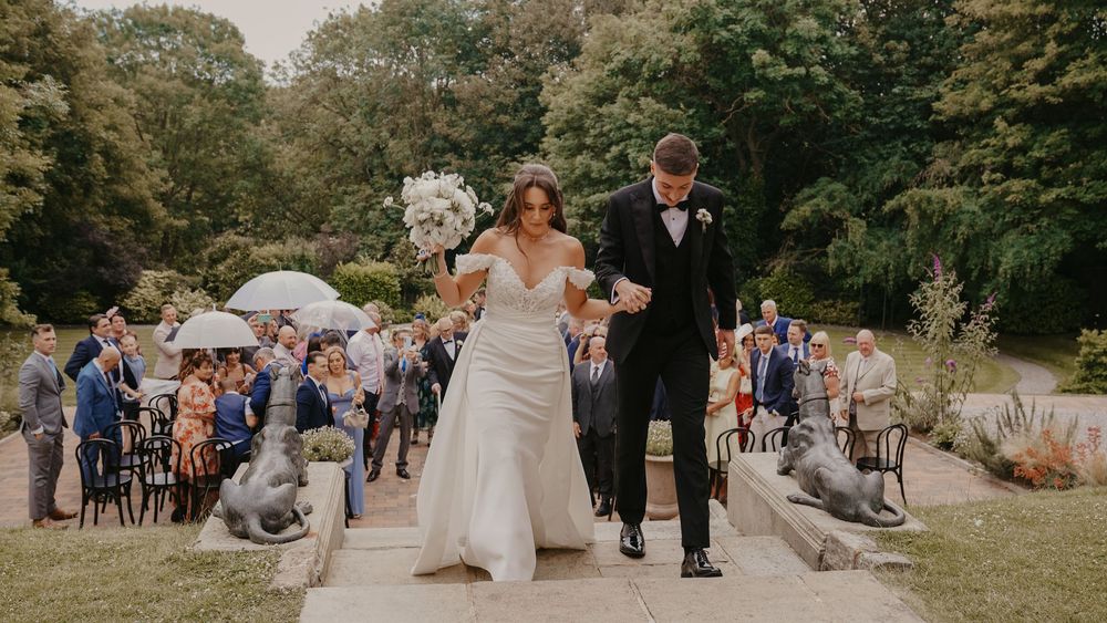 Bride and groom walk hand-in-hand past stone dog statues at an outdoor wedding ceremony with guests in background.
