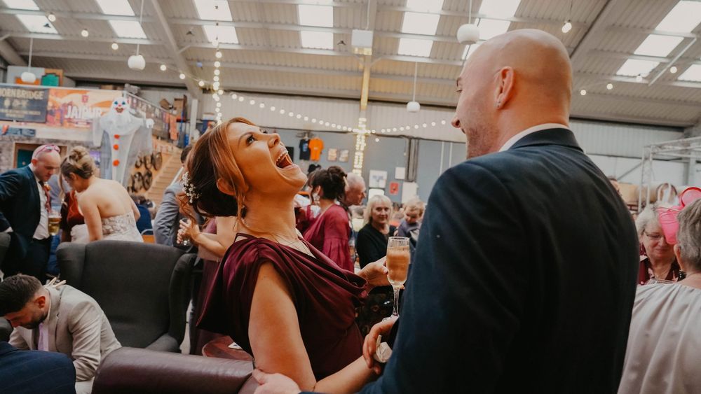 A woman in a burgundy dress laughs joyfully while talking with a bald man in a suit at a lively indoor event.