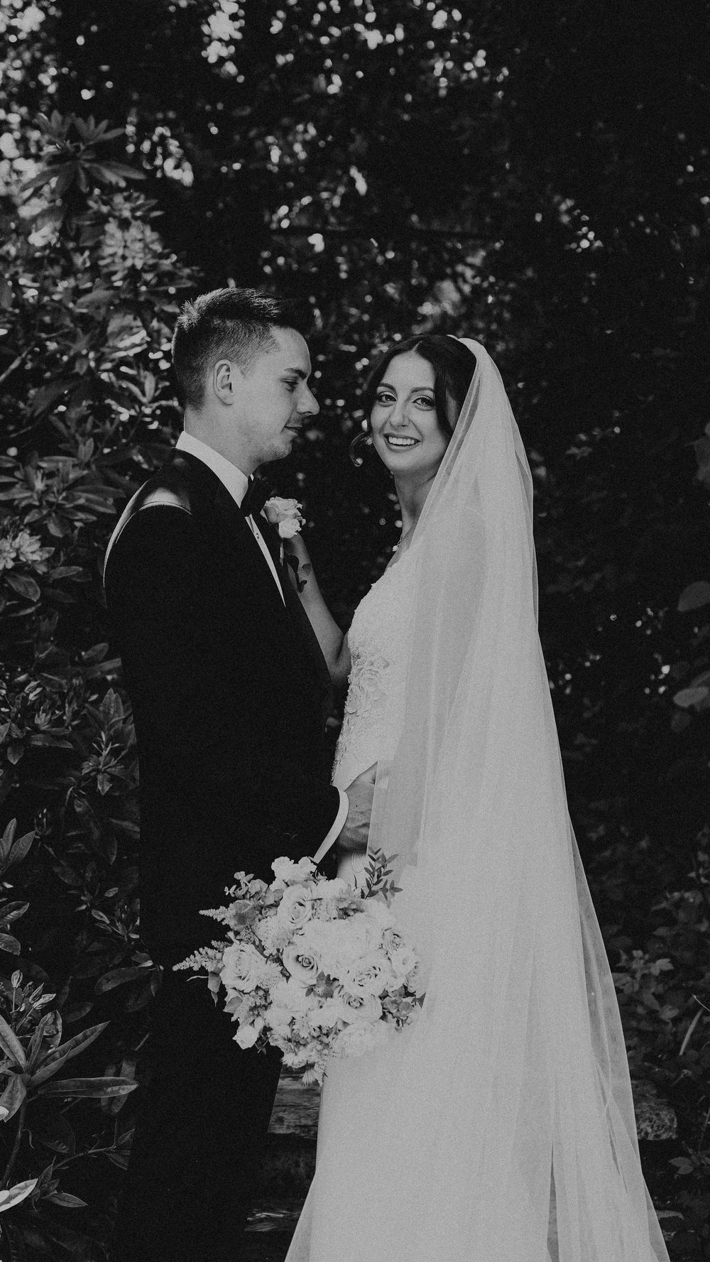 Black and white wedding photo of smiling bride in veil holding bouquet standing with groom in tuxedo outdoors.