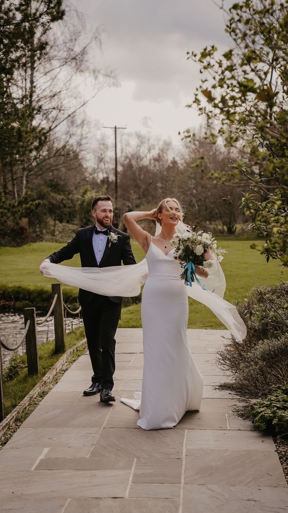 Wedding couple walking on stone path with flowing veil and bouquet in garden setting.