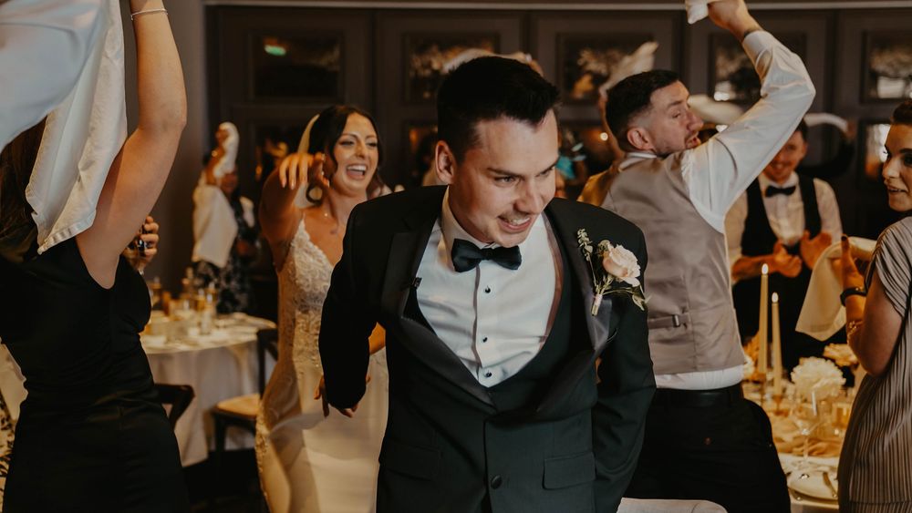 Groom in black tuxedo dancing joyfully with guests at elegant wedding reception celebration.