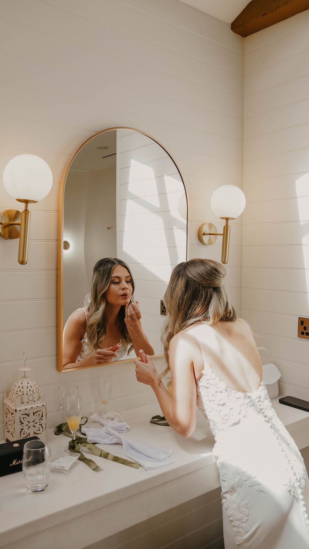 Bride in lace wedding dress applying lipstick at a vanity mirror with gold arch frame and brass wall sconces.