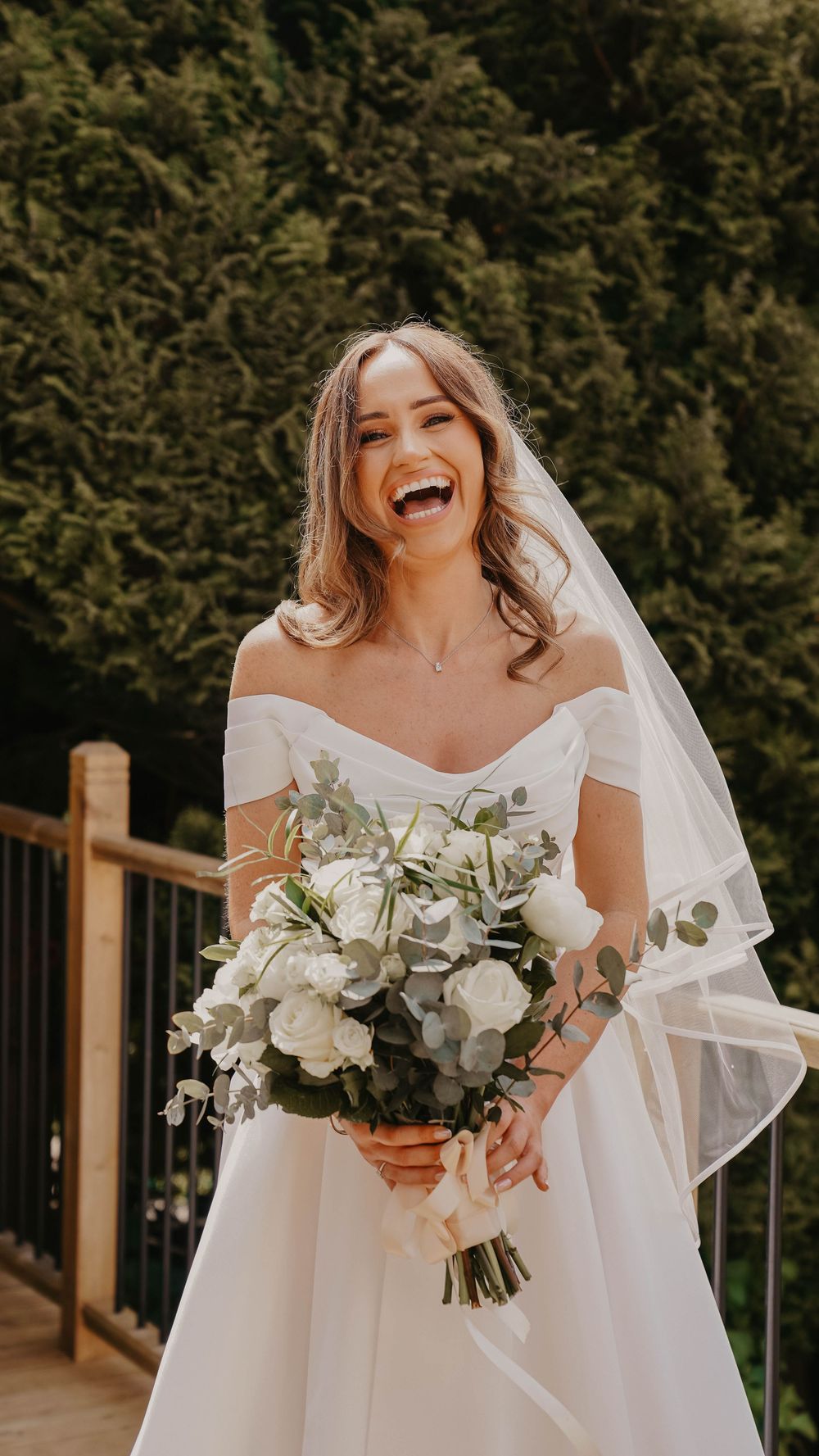 Smiling bride in off-shoulder white gown holding white floral bouquet with veil, standing on wooden deck surrounded by greenery.