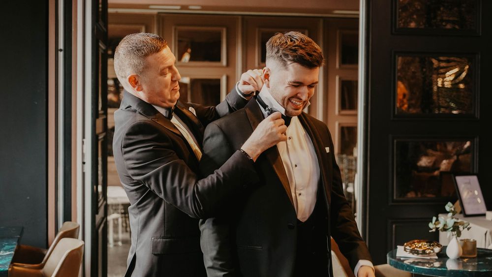Two grooms in tuxedos share a joyful moment as one adjusts the other's boutonniere at a wedding.
