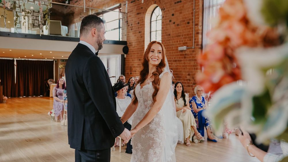 Bride and groom exchange vows at elegant brick wedding venue with large windows and seated guests.