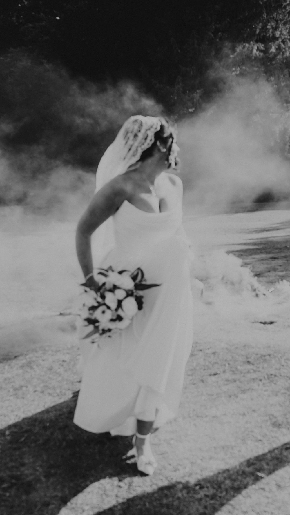Black and white photo of a bride in a white gown and veil holding a bouquet surrounded by dramatic smoke outdoors.