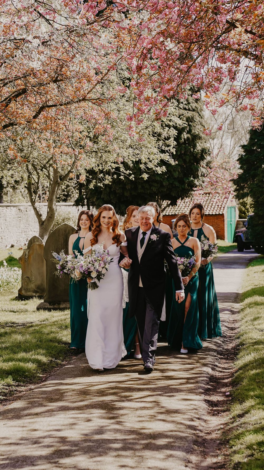 Bride walks with wedding party under blooming cherry blossom trees, bridesmaids in emerald green dresses on sunny spring day.