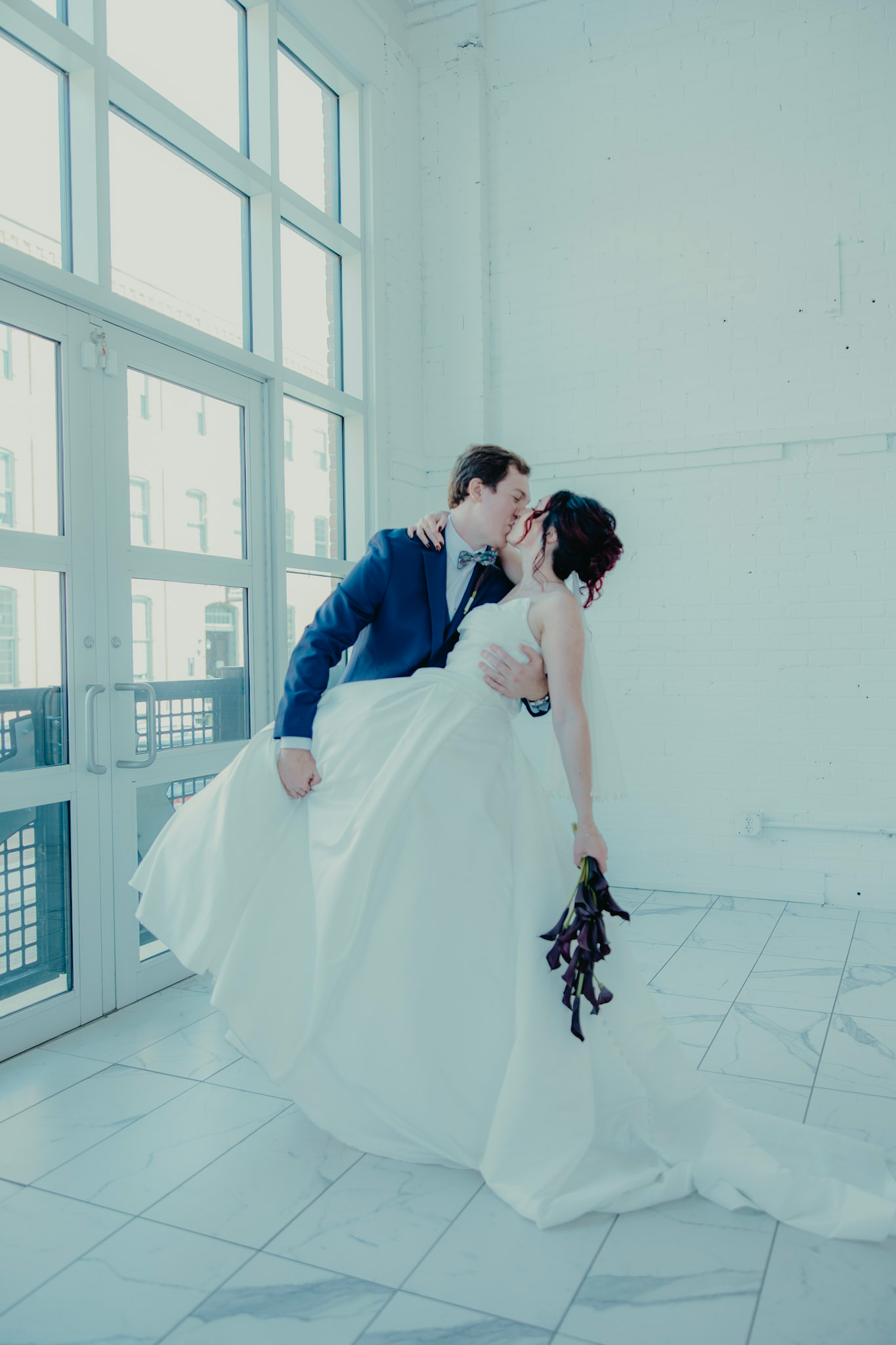 A bride and groom share a romantic dip kiss in a bright white venue, bride holding a dark floral bouquet.