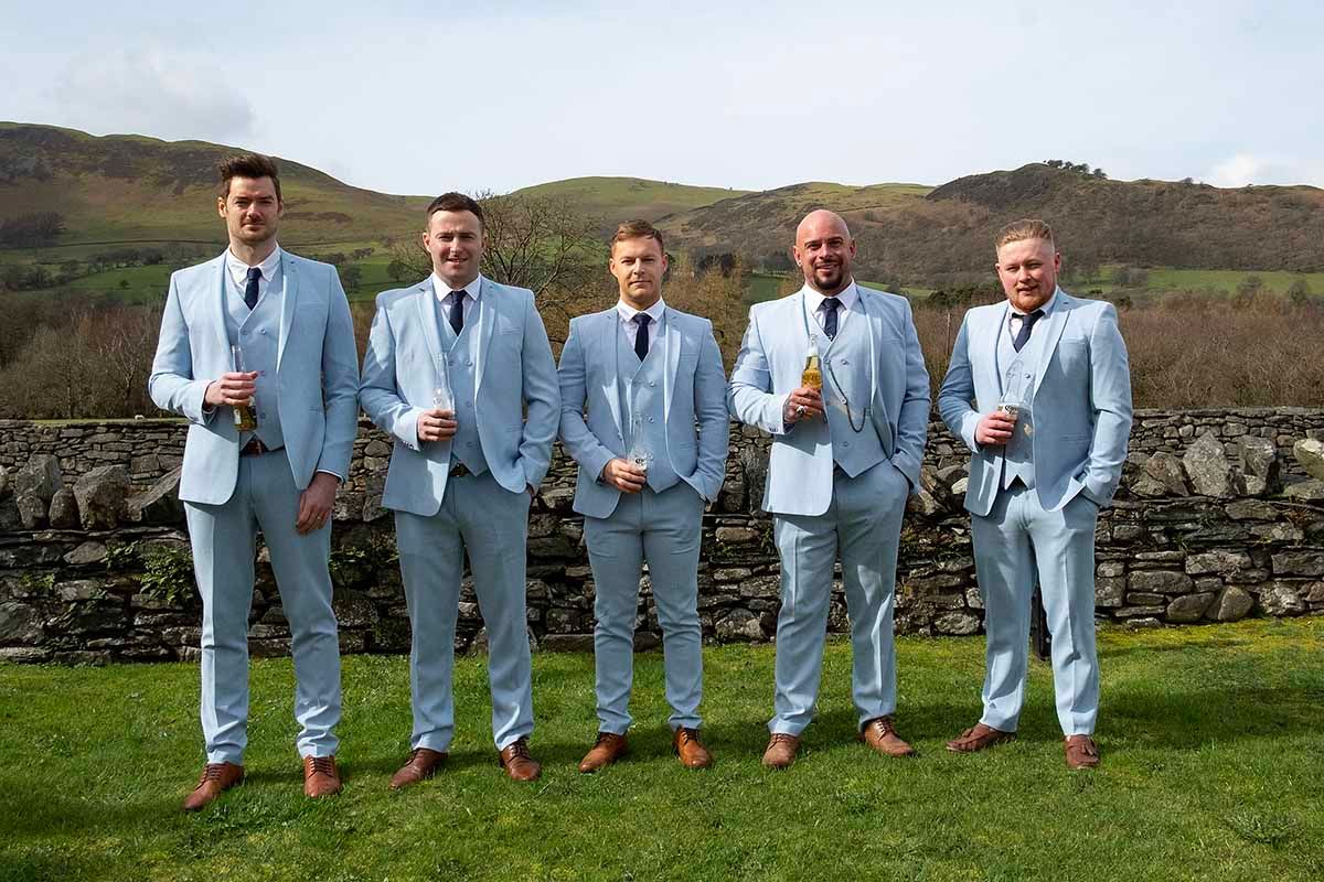 Five groomsmen in matching light blue suits pose outdoors with drinks, rolling green hills of the Lake District at New House Farm