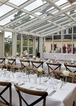 Elegant glass-roofed conservatory dining room with white tablecloths, crystal glasses, white roses, and rustic wooden cross-back chairs.
