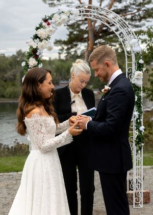 Bride and groom exchange rings under a floral arch outdoors, officiated by a woman, with a lake in the background.