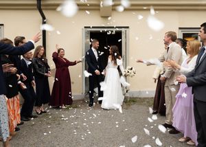 Newlywed couple exits wedding venue as guests toss white confetti petals in joyful outdoor celebration.