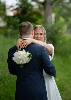 Bride in white dress holding white peonies embraces groom in navy suit outdoors with lush green background.
