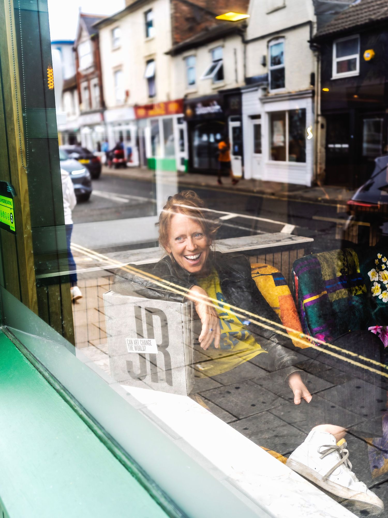 Smiling person reflected in a shop window on a busy UK high street with colorful storefronts visible behind.