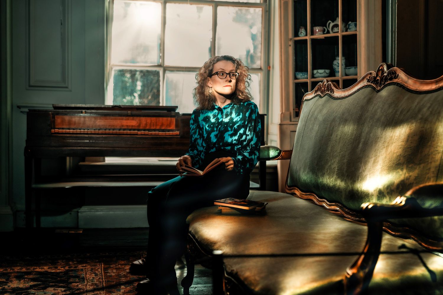 Woman with curly hair reads a book on a vintage settee beside a piano in a warmly lit, elegant room.
