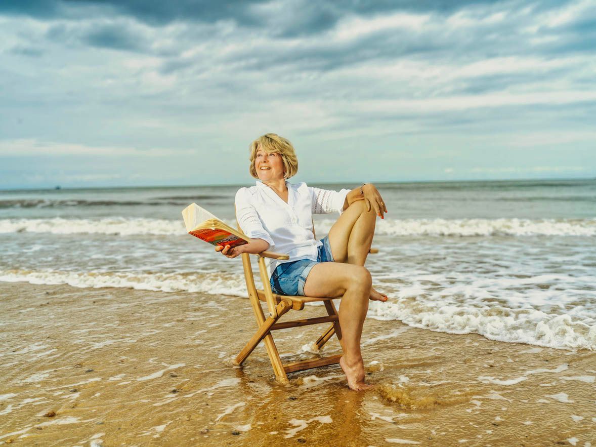 Woman relaxing on a wooden chair at the beach, reading a book with ocean waves in the background.