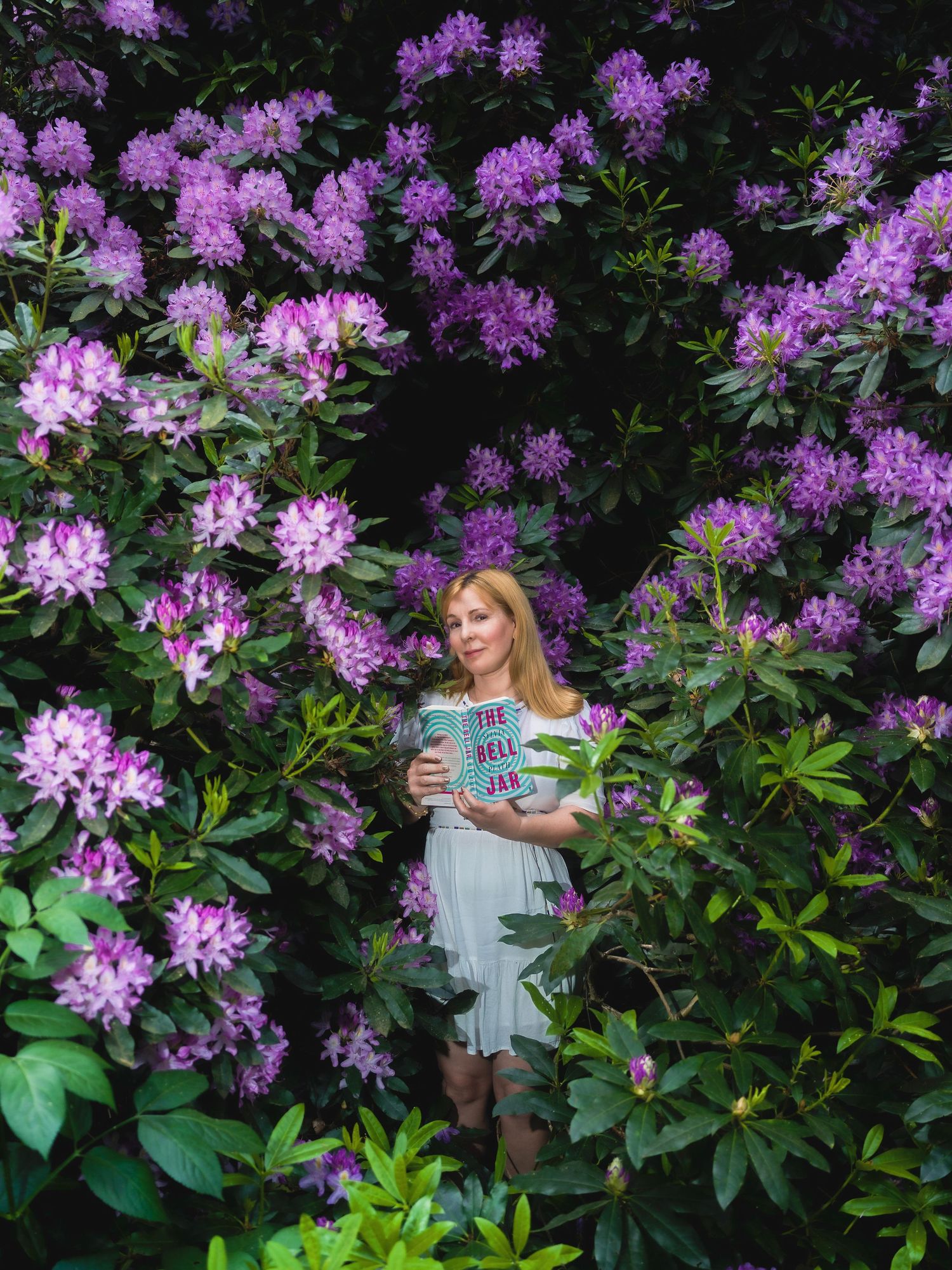 A woman in a light blue dress holds a book surrounded by vibrant purple rhododendron blooms in a lush garden.