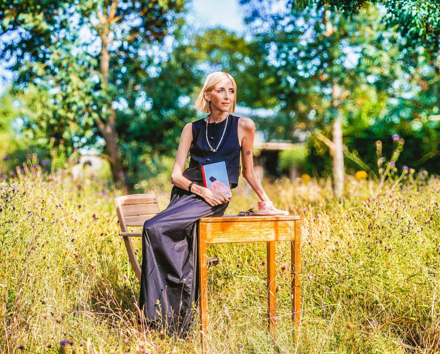 Elegant blonde woman in navy outfit sitting on wooden chair among wildflowers in a lush garden setting.
