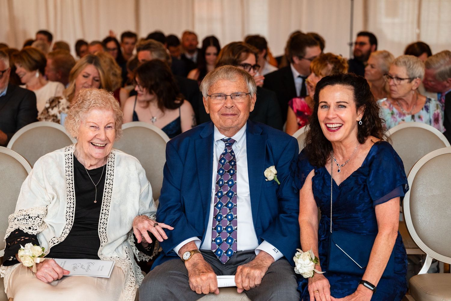 Elderly woman and man in blue suit smile with younger woman at wedding reception seating.