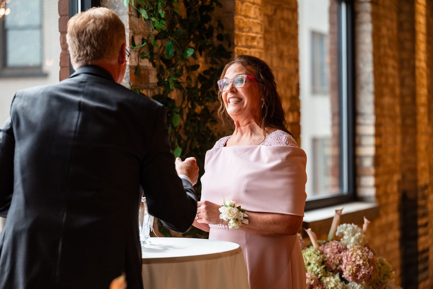Bride in pink dress laughing with groom in black suit during wedding ceremony in rustic brick venue.