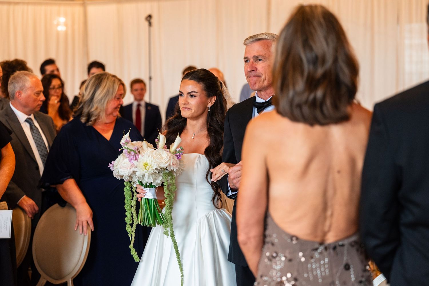 Bride in white gown walks down aisle with parents during elegant wedding ceremony.