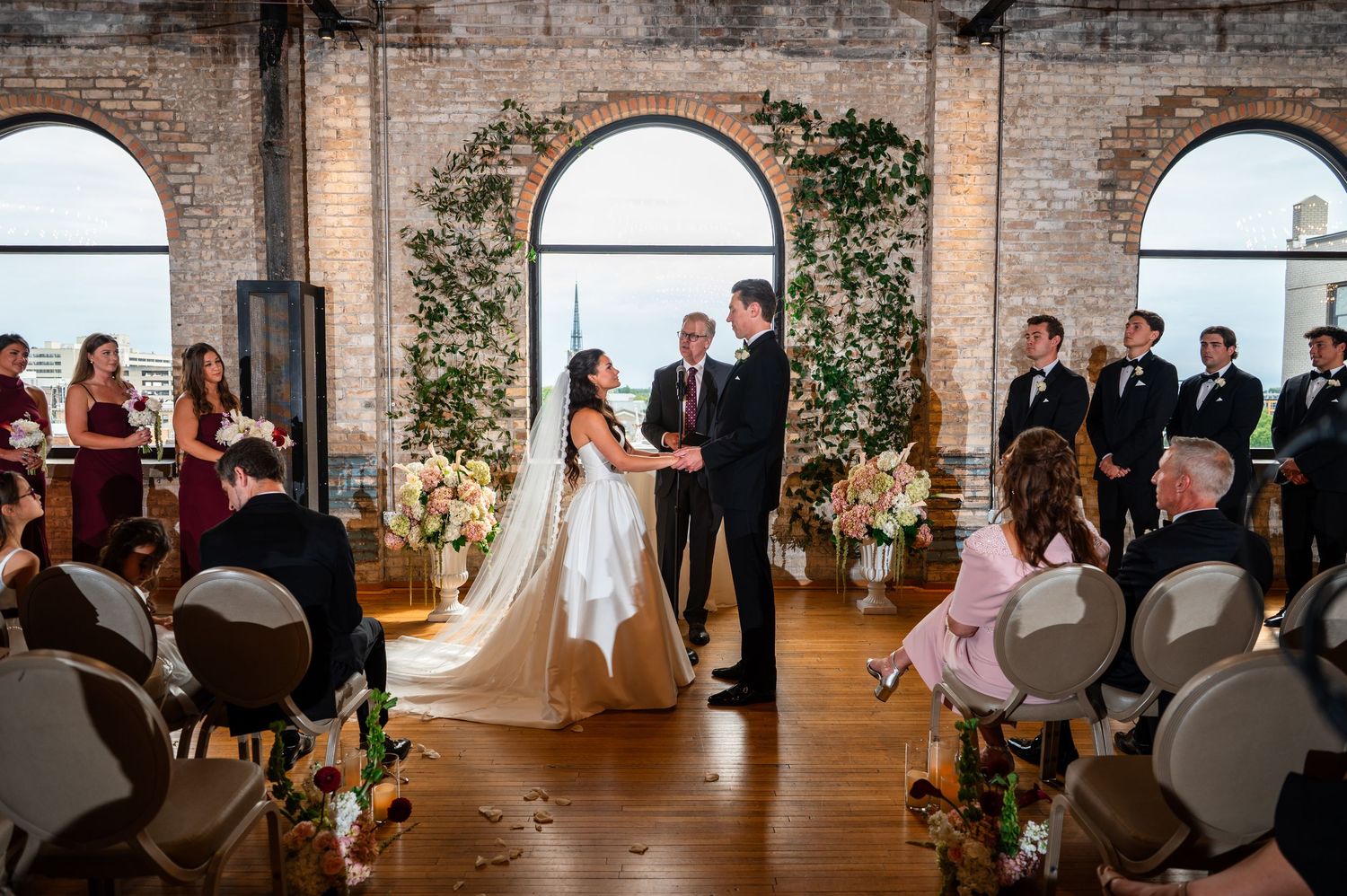Bride and groom exchange vows at a romantic indoor wedding ceremony with lush floral arch and brick walls.