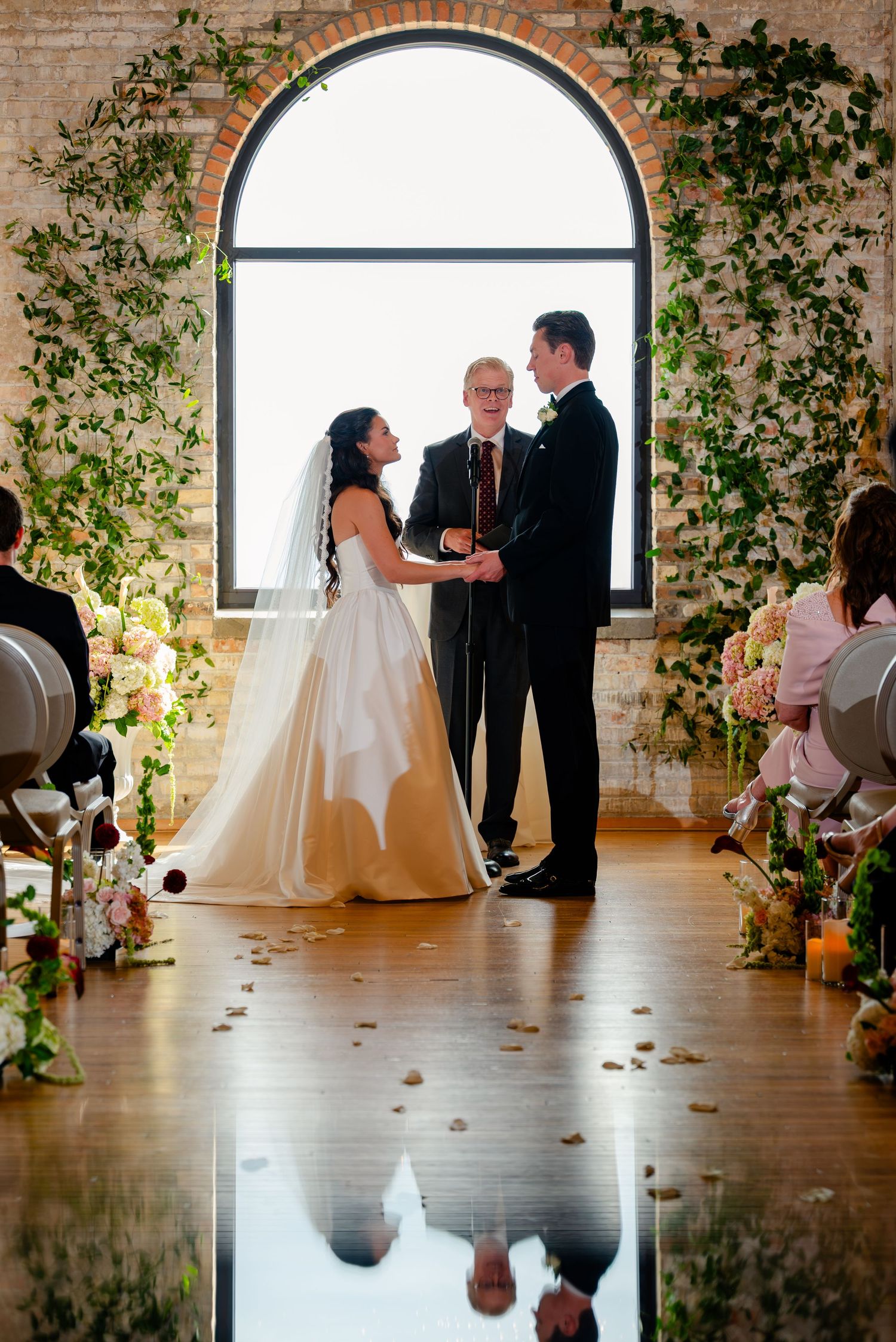 Bride and groom exchange vows before an arched window adorned with lush greenery at an elegant wedding ceremony.