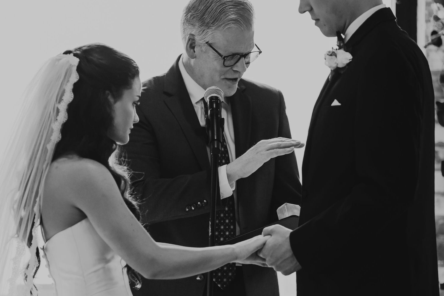 Black and white wedding ceremony photo of bride and groom exchanging rings with officiant.