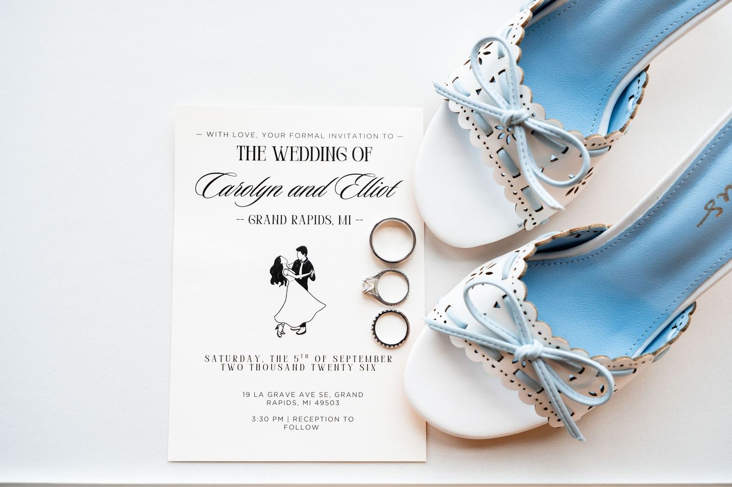 Wedding invitation for Carolyn and Elliot in Grand Rapids, MI, displayed beside elegant light blue bridal flats and gold rings.