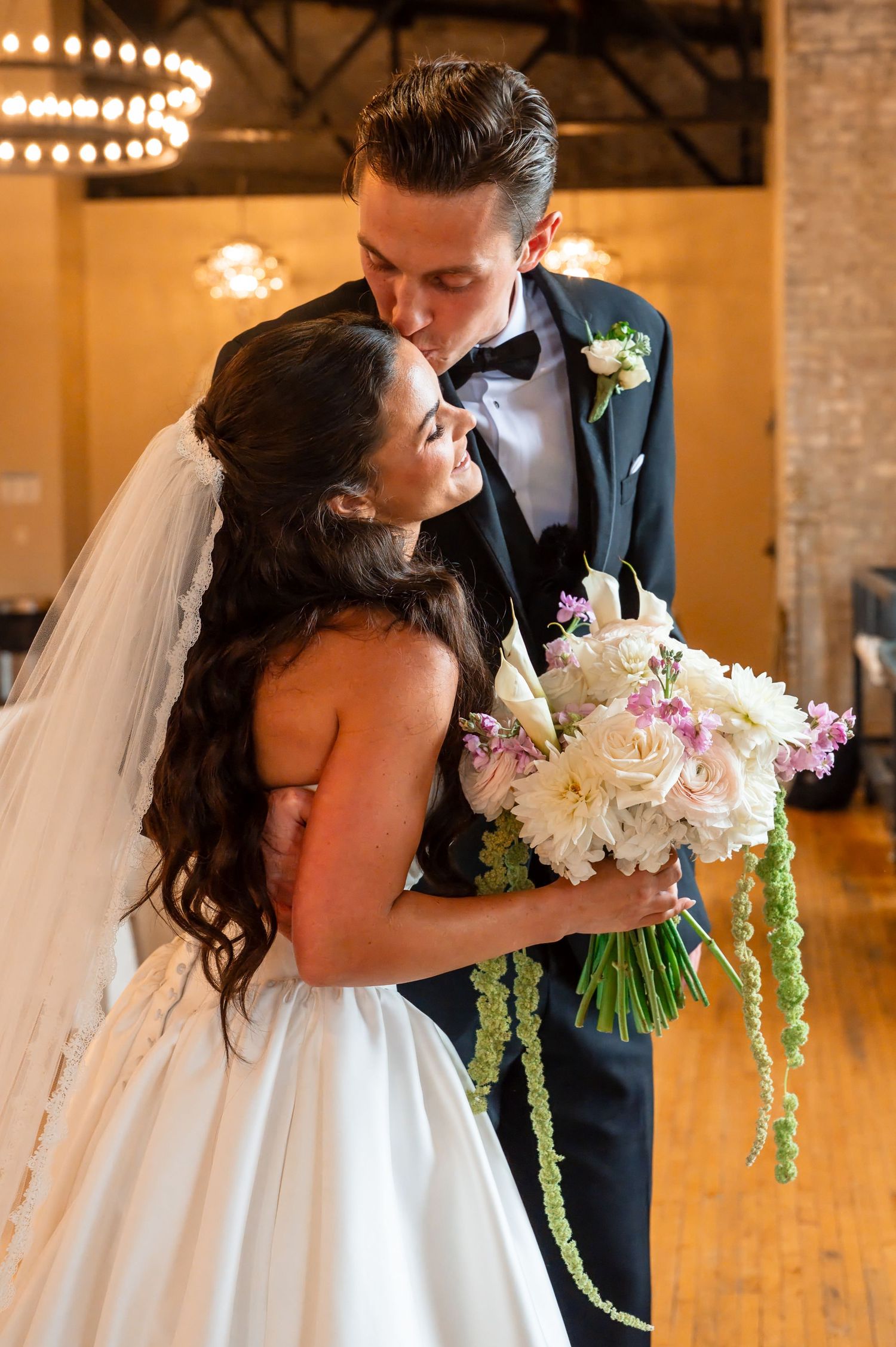 Bride and groom share a romantic kiss, bride holding a lush bouquet of white and pink flowers with green accents.