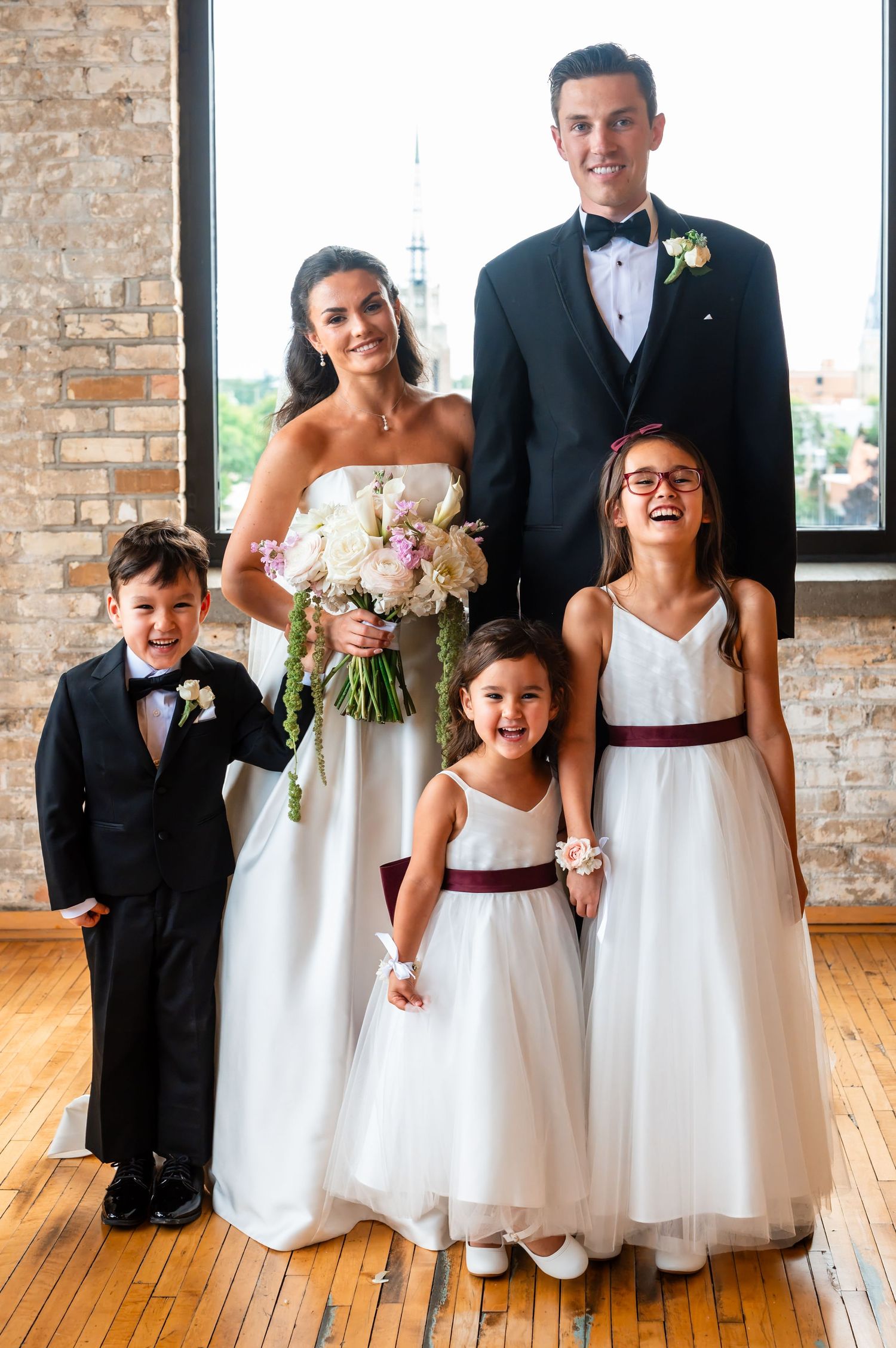 Bride and groom pose with young ring bearer and two flower girls in white dresses with burgundy sashes.