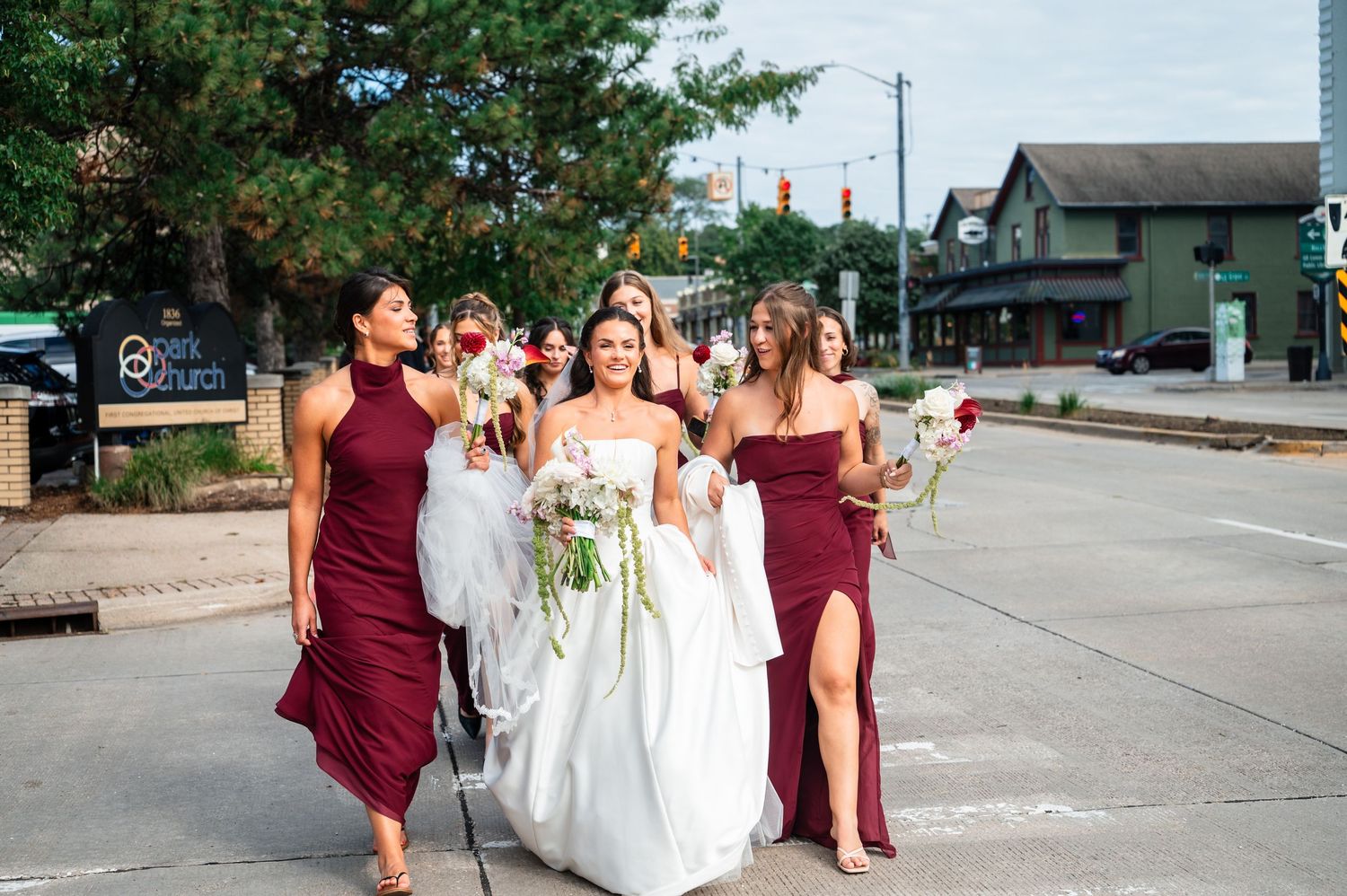 Bride in white gown walks with bridesmaids in burgundy dresses along a city street holding bouquets.