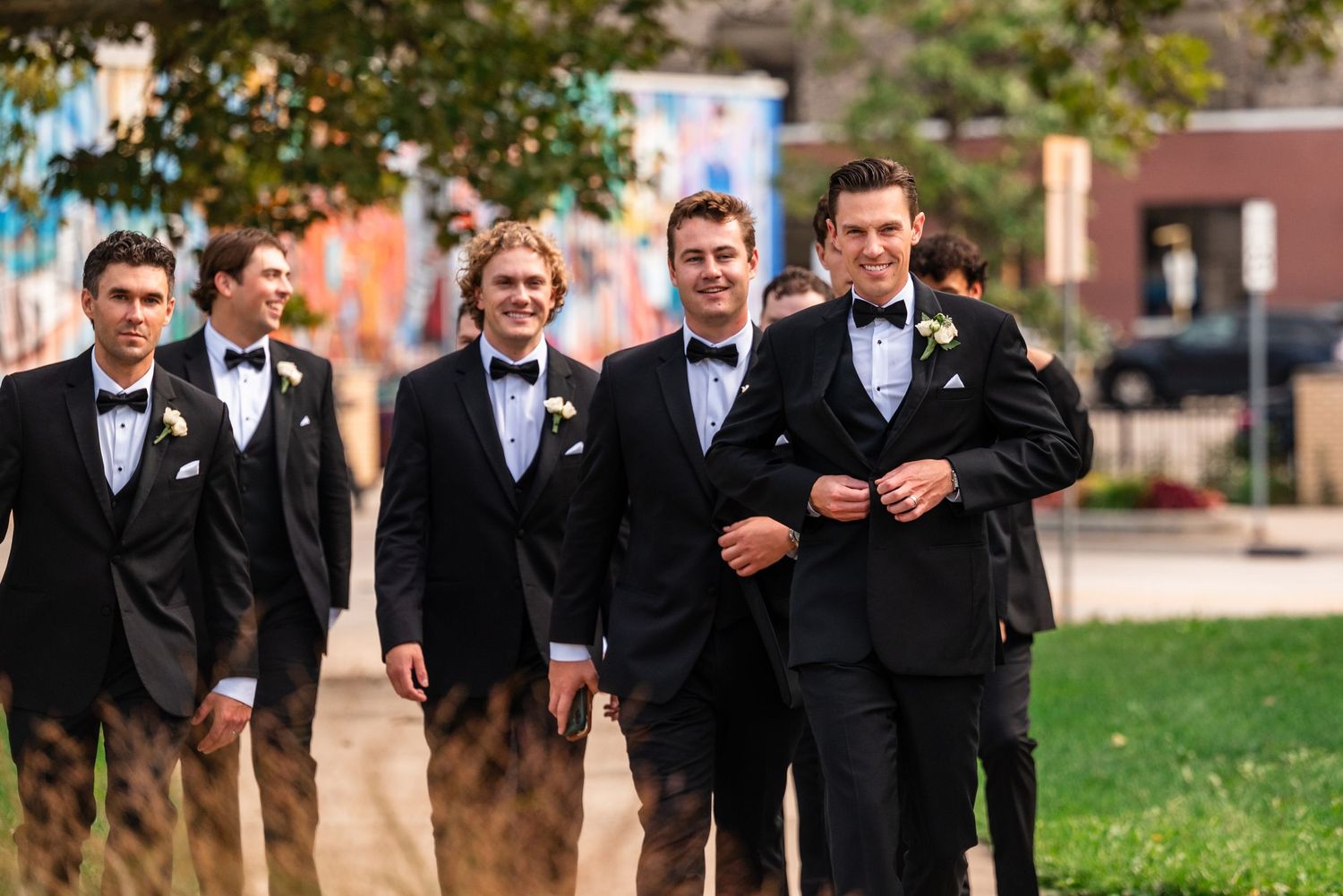 Groomsmen in black tuxedos with bow ties and boutonnieres walk together outdoors before a wedding ceremony.