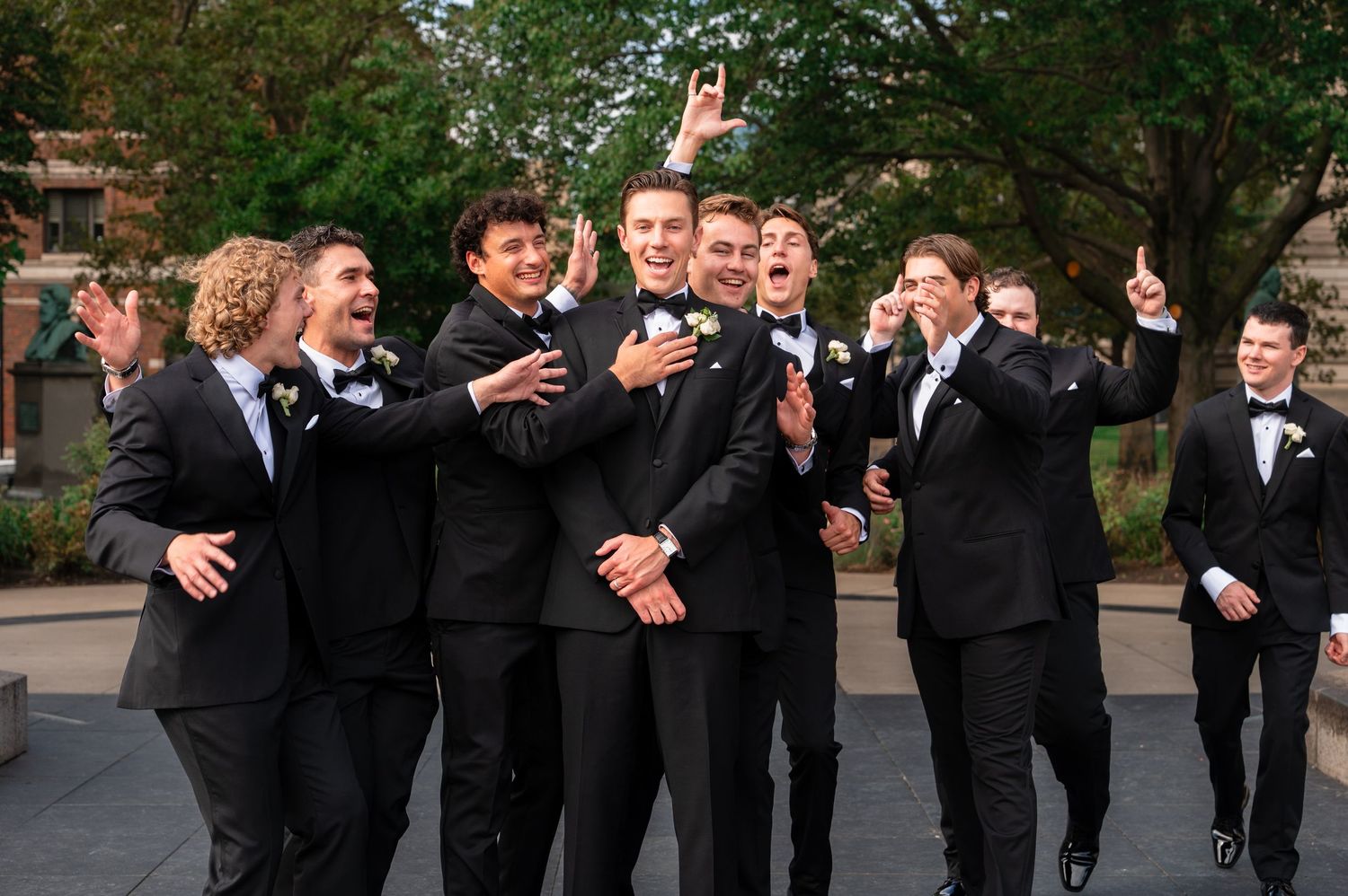 Excited groomsmen in black tuxedos celebrate outdoors with the groom at a wedding party photo.