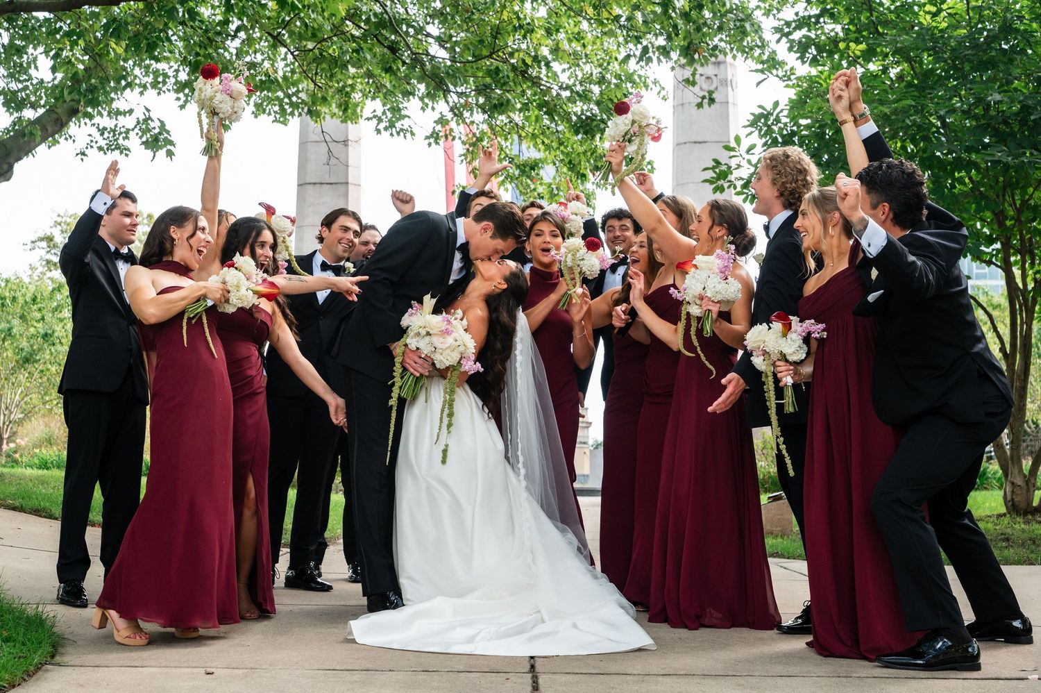 Wedding party celebrating as bride and groom kiss, bridesmaids in burgundy dresses cheering outdoors.