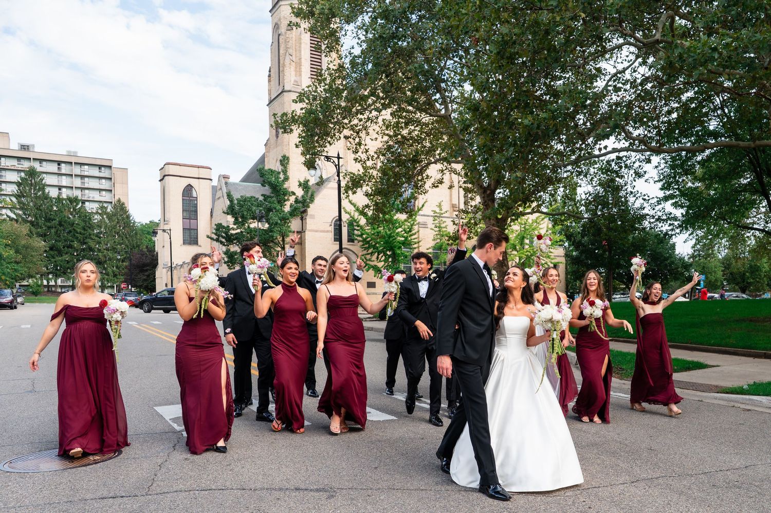 Wedding party in burgundy and black attire walks across street near gothic church during outdoor photos.