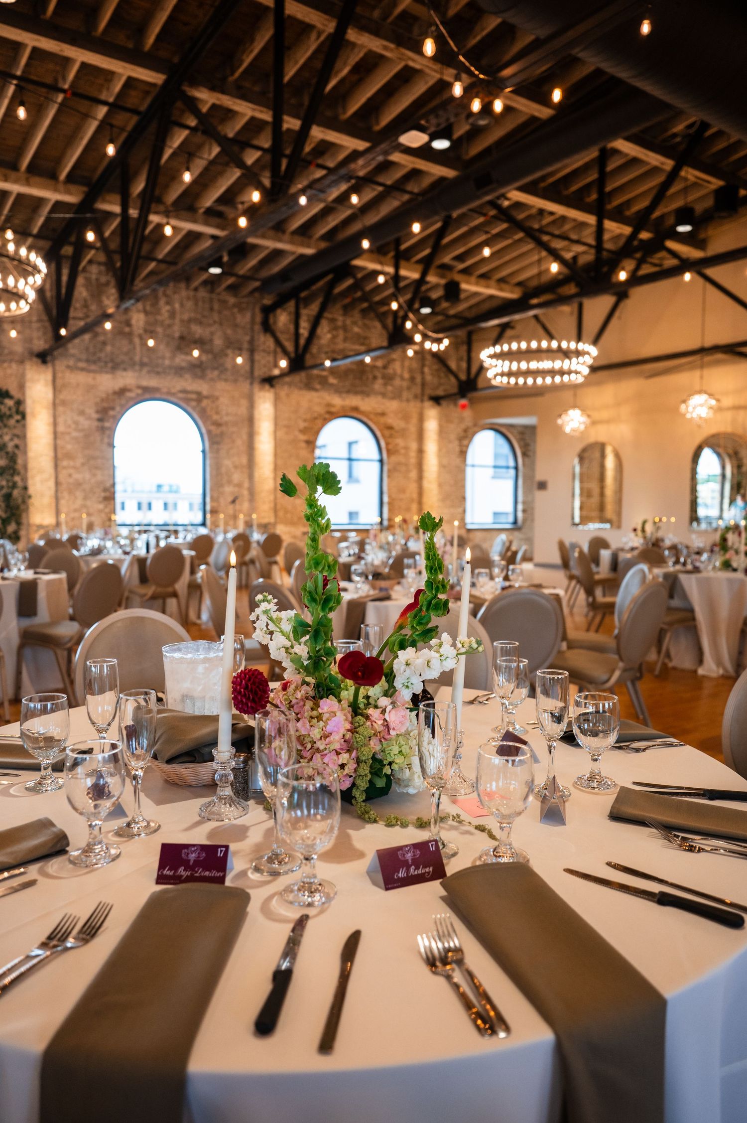 Elegant wedding reception tables with floral centerpieces of burgundy and pink blooms in brick ballroom.