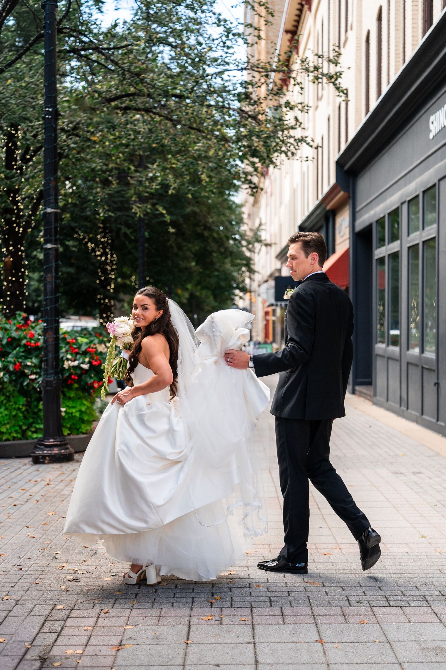Bride in white gown and groom in black suit walk together on a city sidewalk during wedding photos.