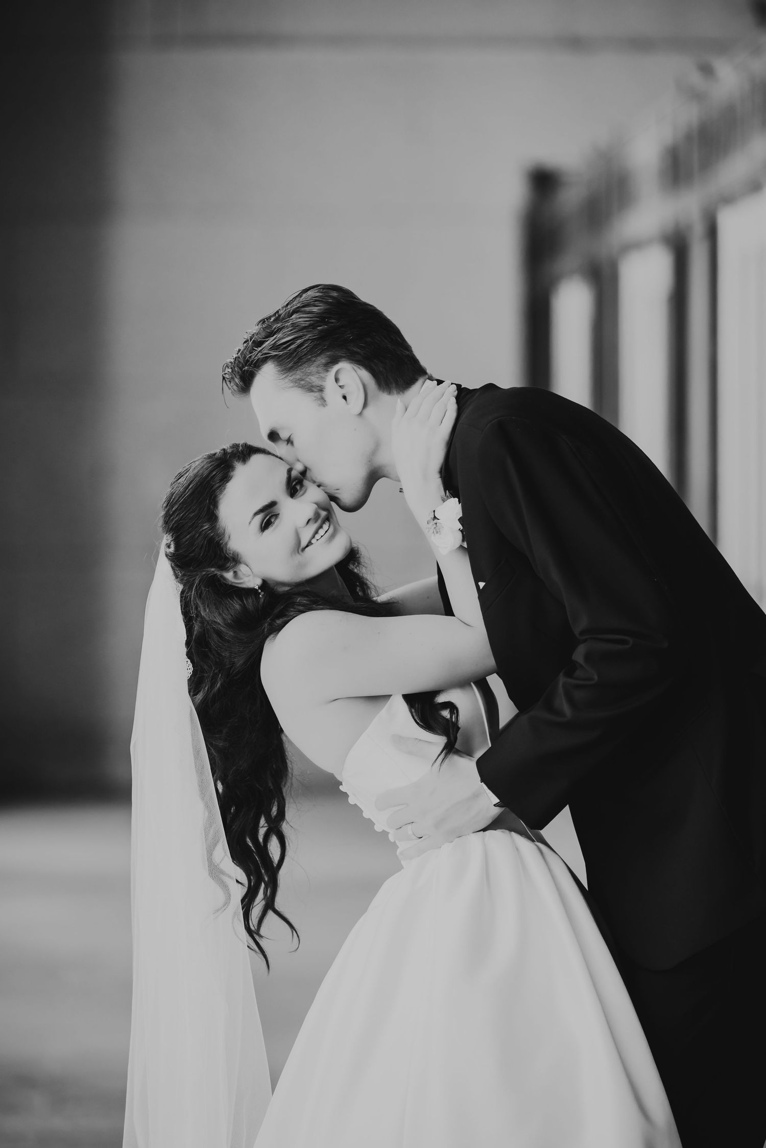 Black and white wedding photo of smiling bride in veil and groom in suit sharing a romantic kiss.