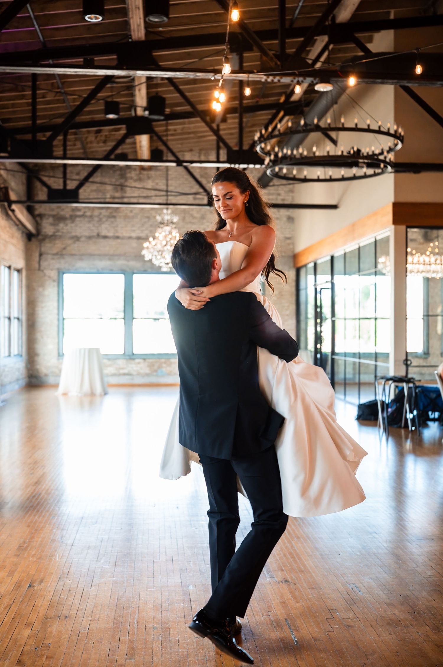 Groom lifting bride during first dance in elegant wedding venue with exposed brick walls and chandelier lighting.
